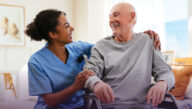 A smiling nurse crouches down and puts her arms around an older man in a wheelchair.